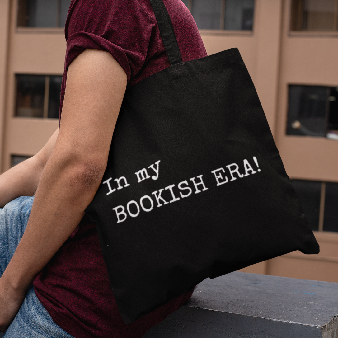 Man wearing black tote bag with “In my BOOKISH ERA!” text, sitting outdoors in an urban setting