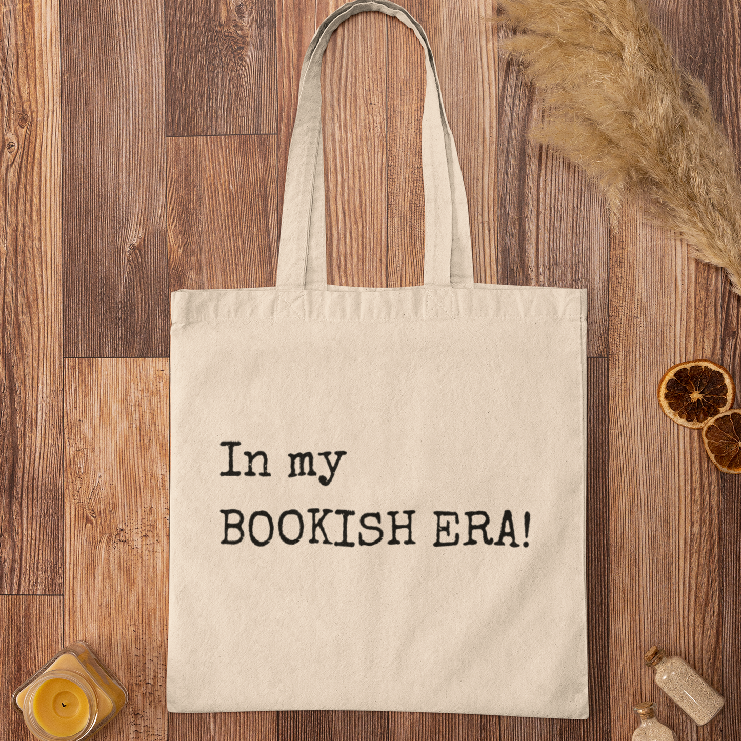 Minimalist tote bag with “In my BOOKISH ERA!” quote printed in typewriter font, displayed on a wooden surface with rustic décor.