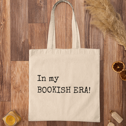 Minimalist tote bag with “In my BOOKISH ERA!” quote printed in typewriter font, displayed on a wooden surface with rustic décor.