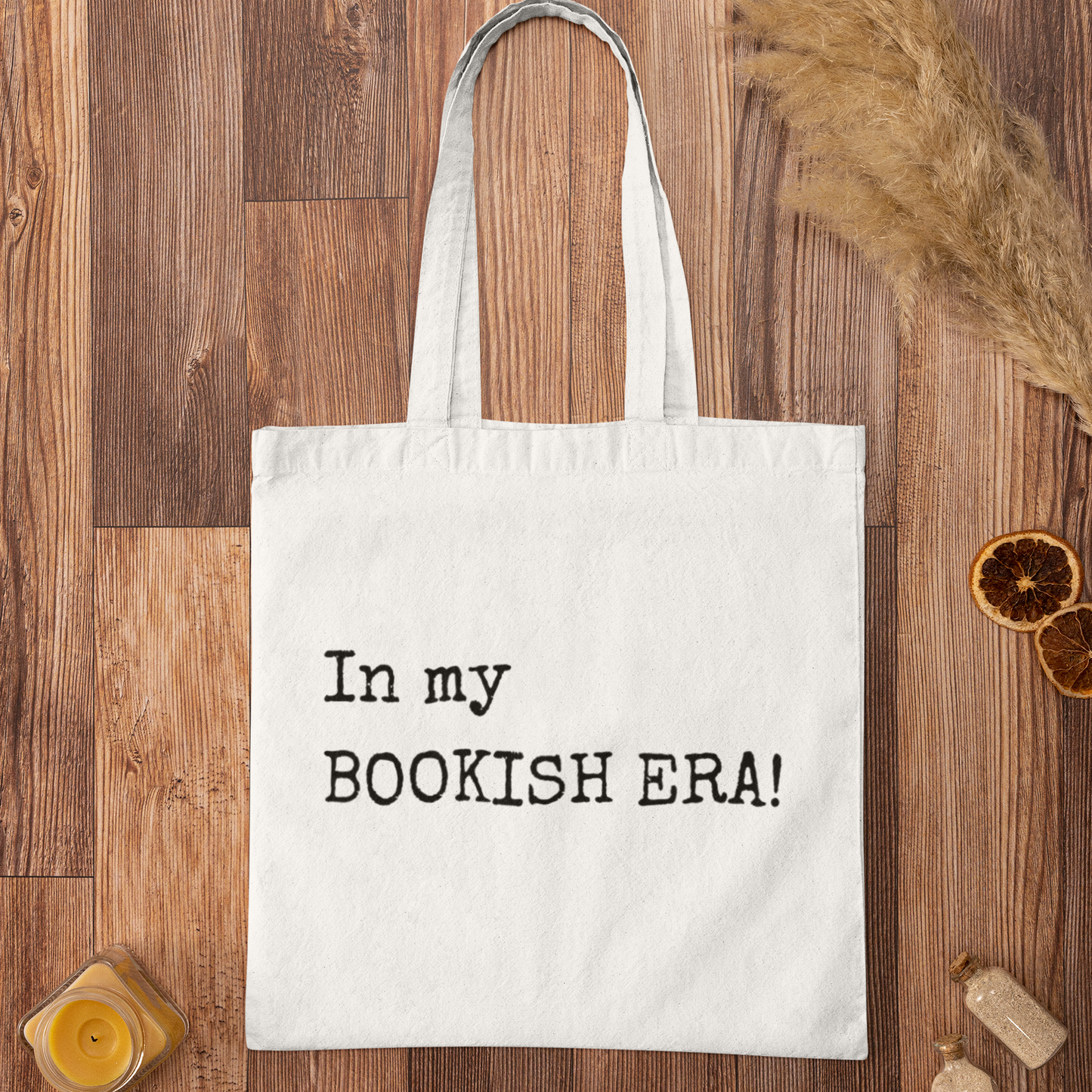 White version of “In my BOOKISH ERA!” minimalist tote bag displayed on a warm wood background with pampas grass accent.