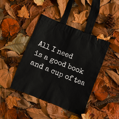 Black book quote tote bag reading 'All I need is a good book and a cup of tea' placed on autumn leaves – cozy fall aesthetic tote bag for readers