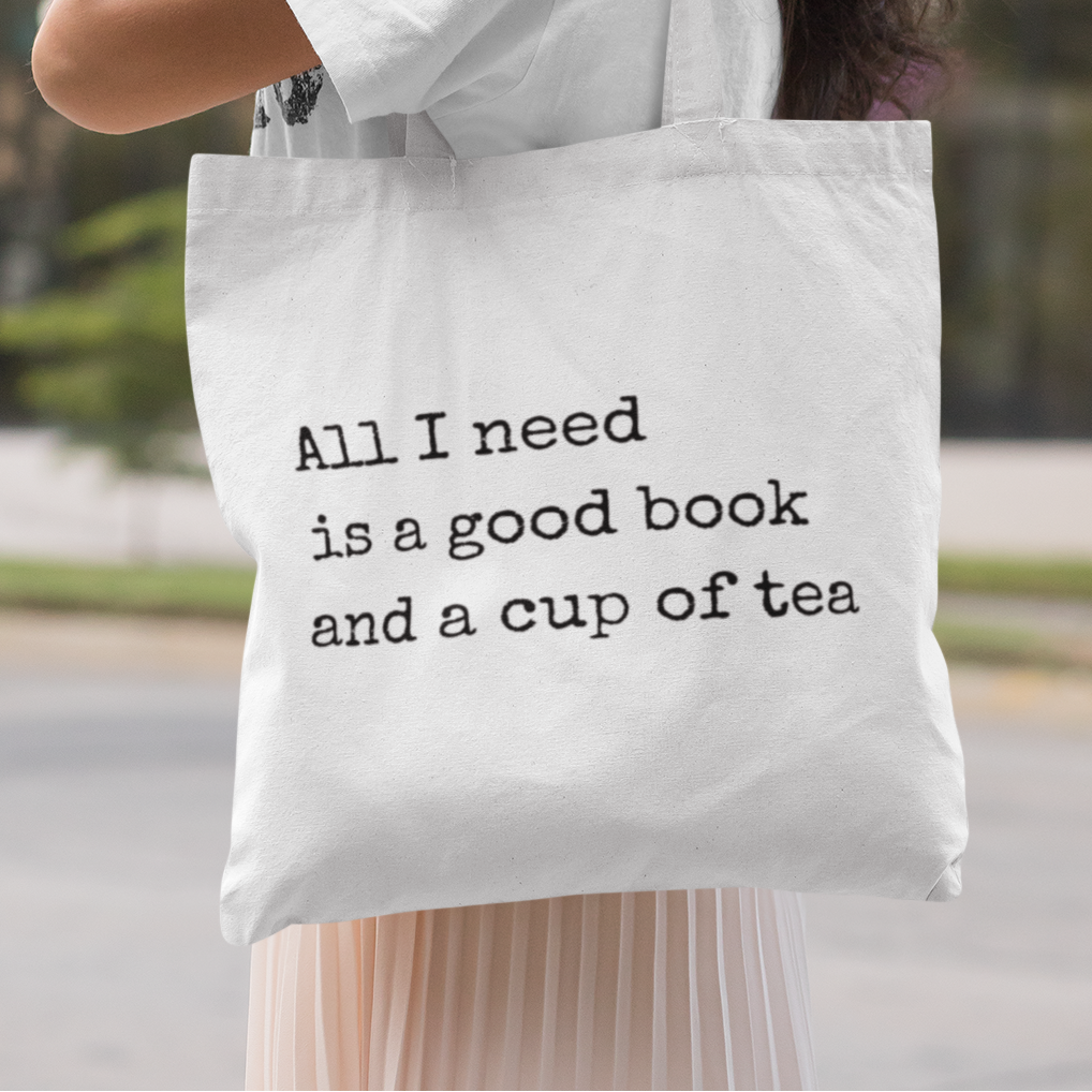 Smiling woman wearing white t-shirt and headphones carrying a white tote bag with quote 'All I need is a good book and a cup of tea' – minimalist book lover tote bag