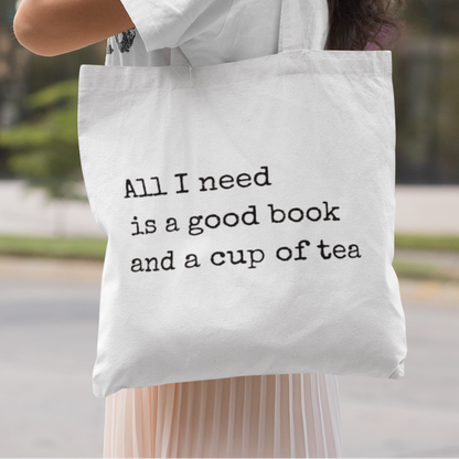 Smiling woman wearing white t-shirt and headphones carrying a white tote bag with quote 'All I need is a good book and a cup of tea' – minimalist book lover tote bag