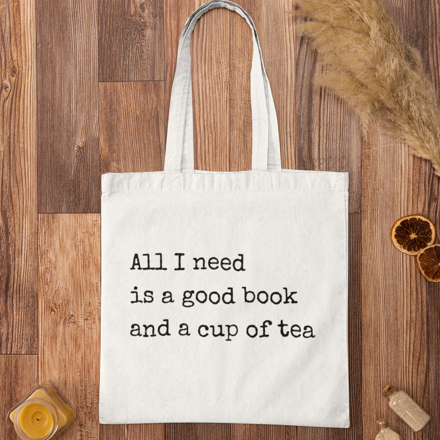 White minimalist tote bag with printed quote 'All I need is a good book and a cup of tea' displayed on wooden background – aesthetic bookish accessory