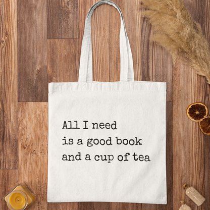 White minimalist tote bag with printed quote 'All I need is a good book and a cup of tea' displayed on wooden background – aesthetic bookish accessory