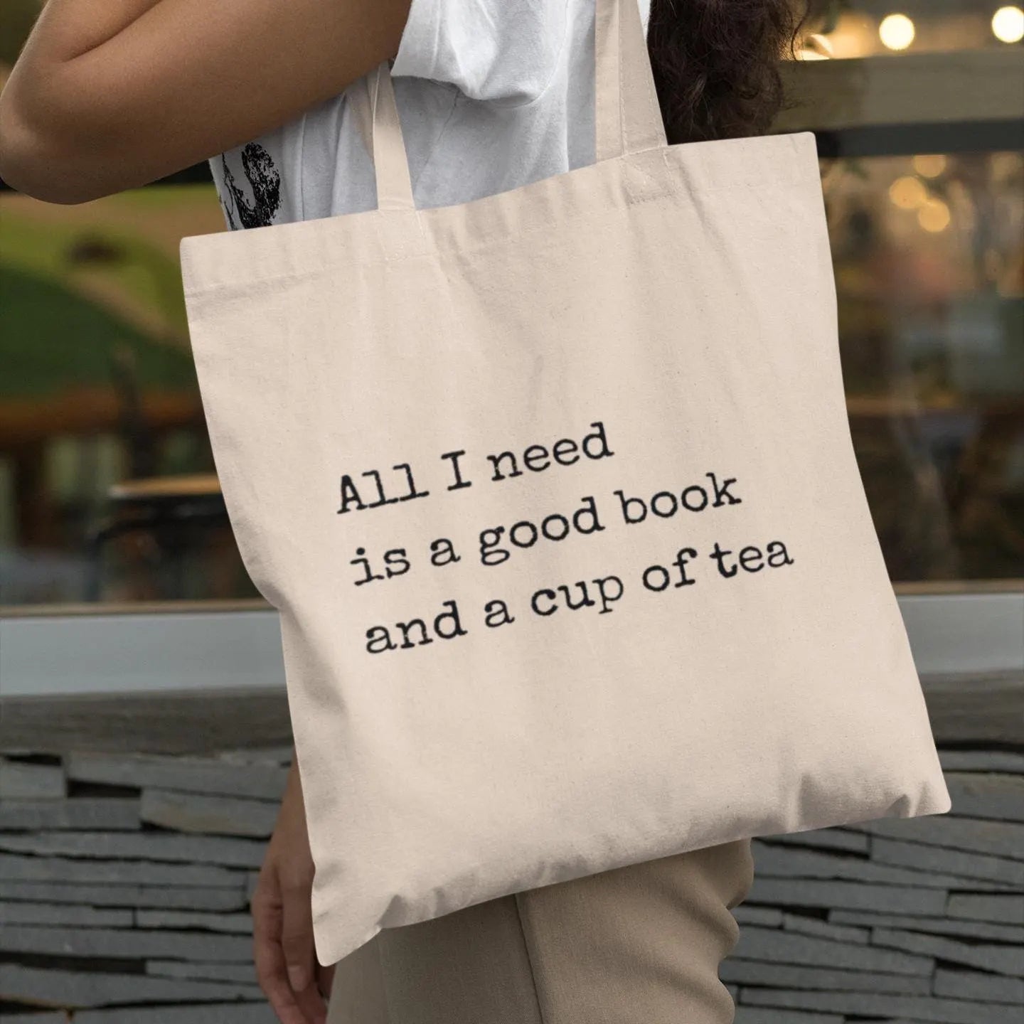 Close-up of woman holding beige canvas tote bag with printed quote 'All I need is a good book and a cup of tea' – minimalist eco tote for book lovers