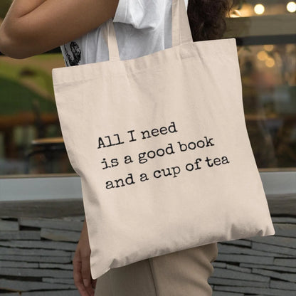 Close-up of woman holding beige canvas tote bag with printed quote 'All I need is a good book and a cup of tea' – minimalist eco tote for book lovers