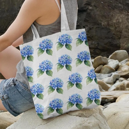 Person sitting on rocks near the beach holding Hydrangea Patterned Floral Design Tote Bag with blue hydrangea pattern