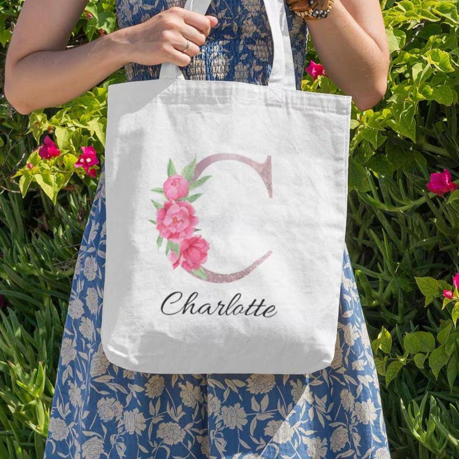 Woman holding white tote bag with blush pink floral initial C and name Charlotte in outdoor garden setting
