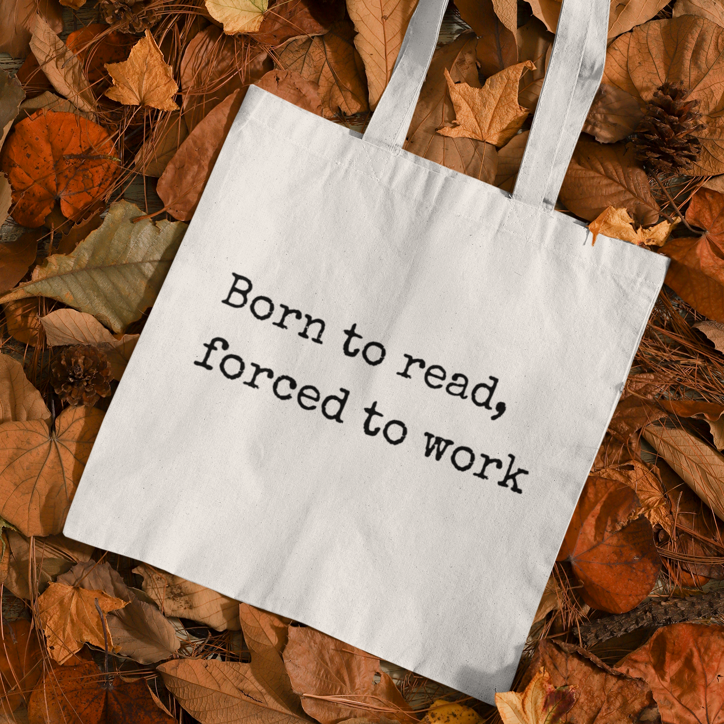 White “Born to read, forced to work” tote bag on autumn leaves — cozy book tote aesthetic