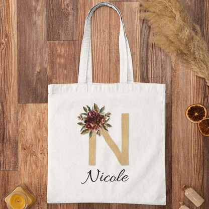 Flat lay of personalized tote bag with gold initial N and burgundy floral print on wooden background