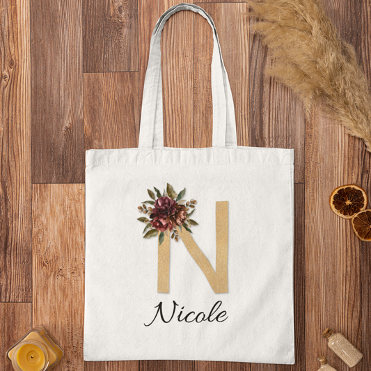 Flat lay of personalized tote bag with gold initial N and burgundy floral print on wooden background