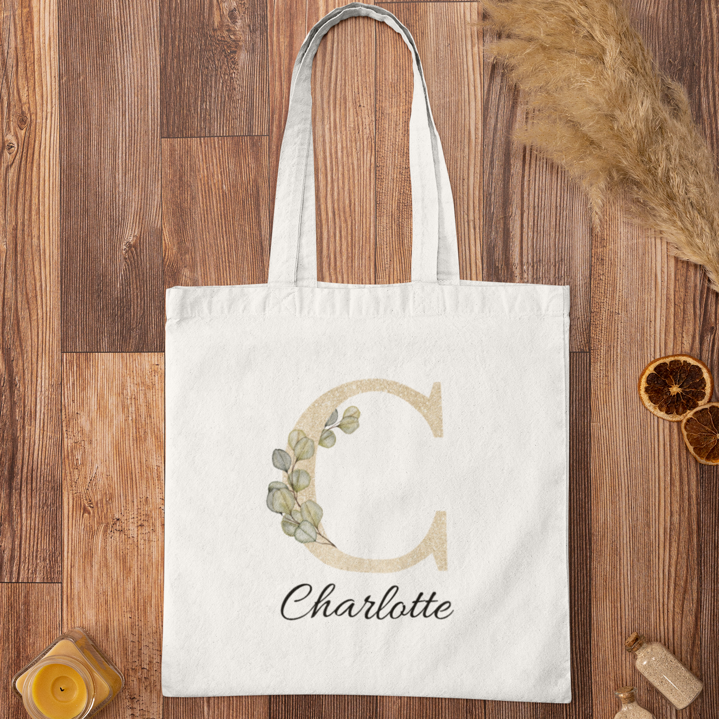 Flat lay of champagne gold and sage green eucalyptus initial tote bag with name Charlotte, personalized cotton tote on wooden background.