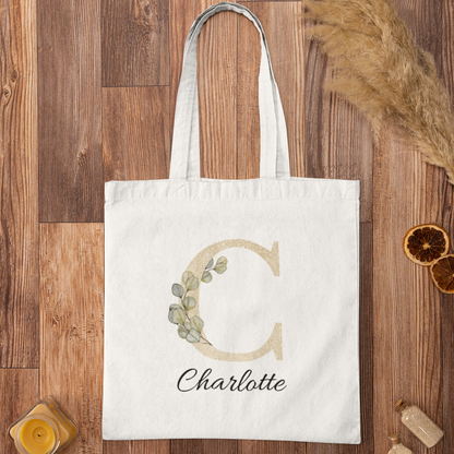 Flat lay of champagne gold and sage green eucalyptus initial tote bag with name Charlotte, personalized cotton tote on wooden background.