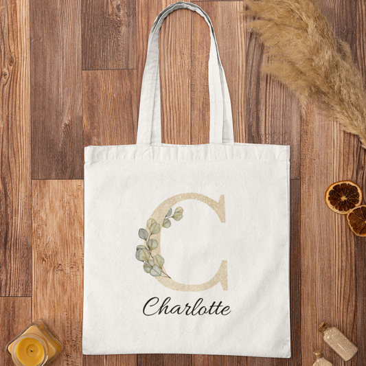 Flat lay of champagne gold and sage green eucalyptus initial tote bag with name Charlotte, personalized cotton tote on wooden background.