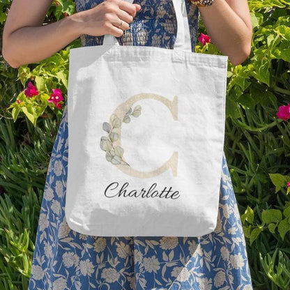 Champagne gold eucalyptus initial tote bag personalized with name Charlotte, photographed outdoors among greenery