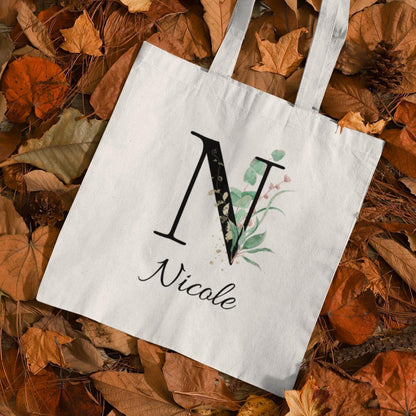 Nicole personalized tote bag with Eucalyptus, Gold, and Berry Leaf initial design displayed on rustic flat lay background