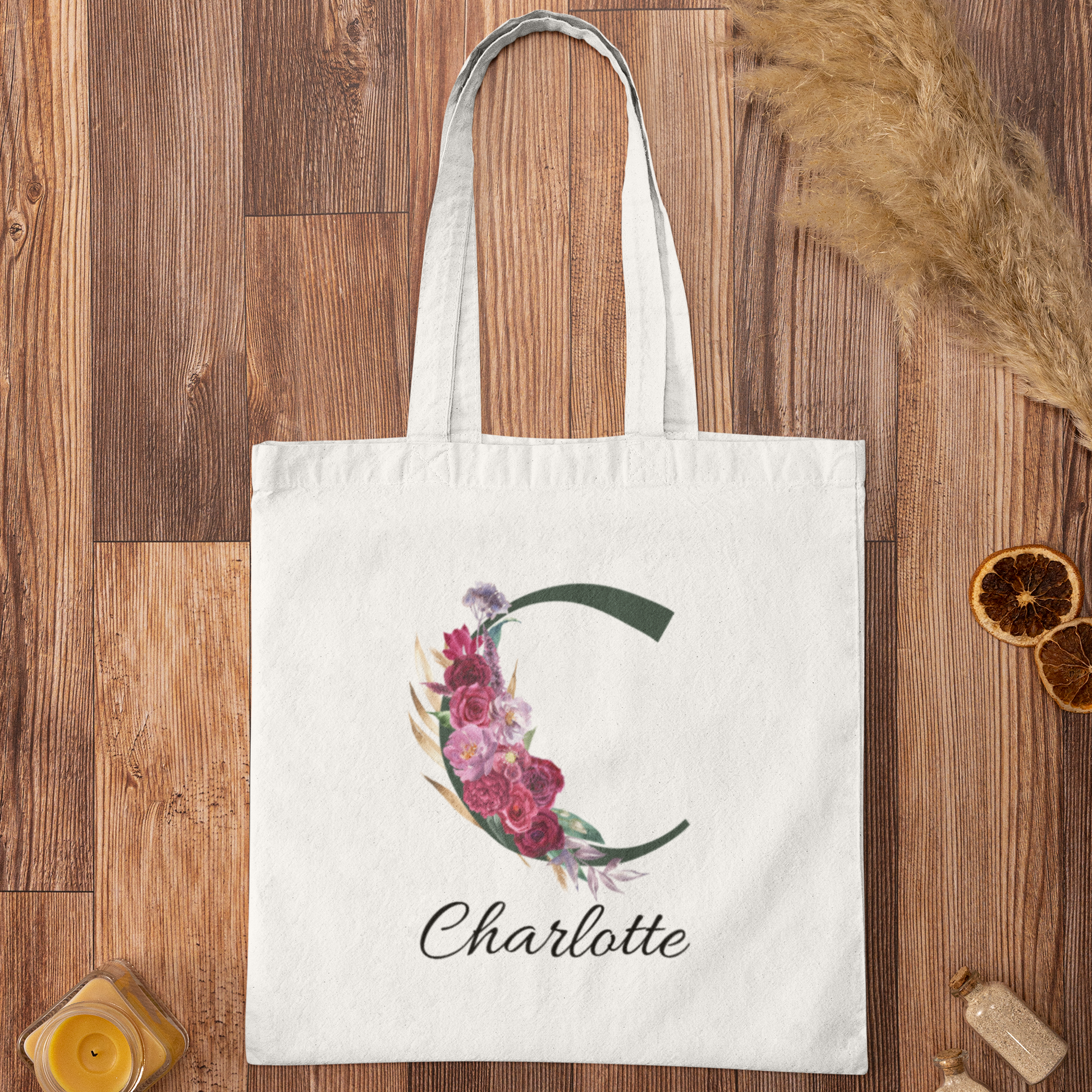 Flatlay of personalised tote bag with letter C and maroon floral design on wooden background with decor items