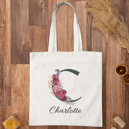 Flatlay of personalised tote bag with letter C and maroon floral design on wooden background with decor items