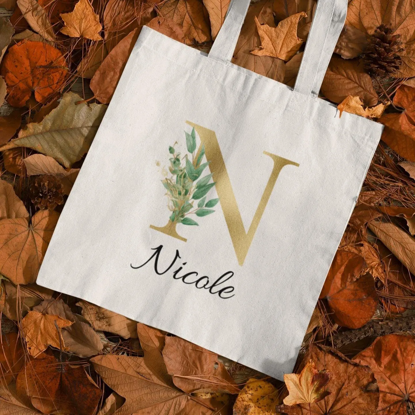 Gold and sage green eucalyptus initial tote bag with letter N and name Nicole, laid on autumn leaves background