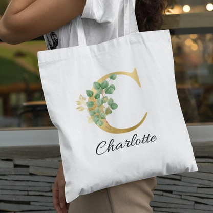 Custom tote bag with gold letter C and eucalyptus leaf design, personalised with name Charlotte, carried by woman outdoors