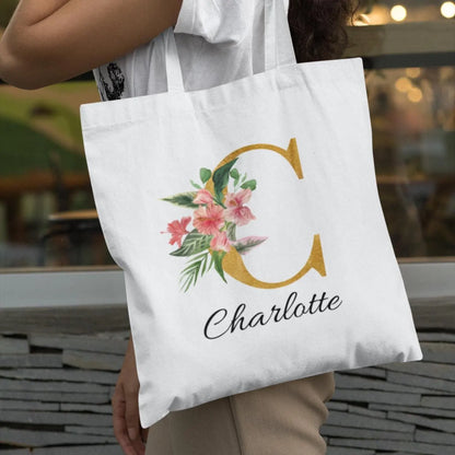 Woman carrying hibiscus tropical floral custom tote bag with gold initial C and name Charlotte, outdoor lifestyle scene