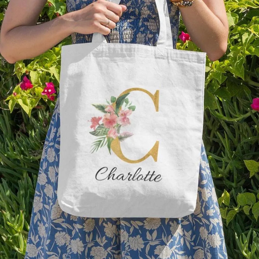 Hibiscus tropical floral custom tote bag with gold initial C and name Charlotte, held outdoors in a floral garden setting