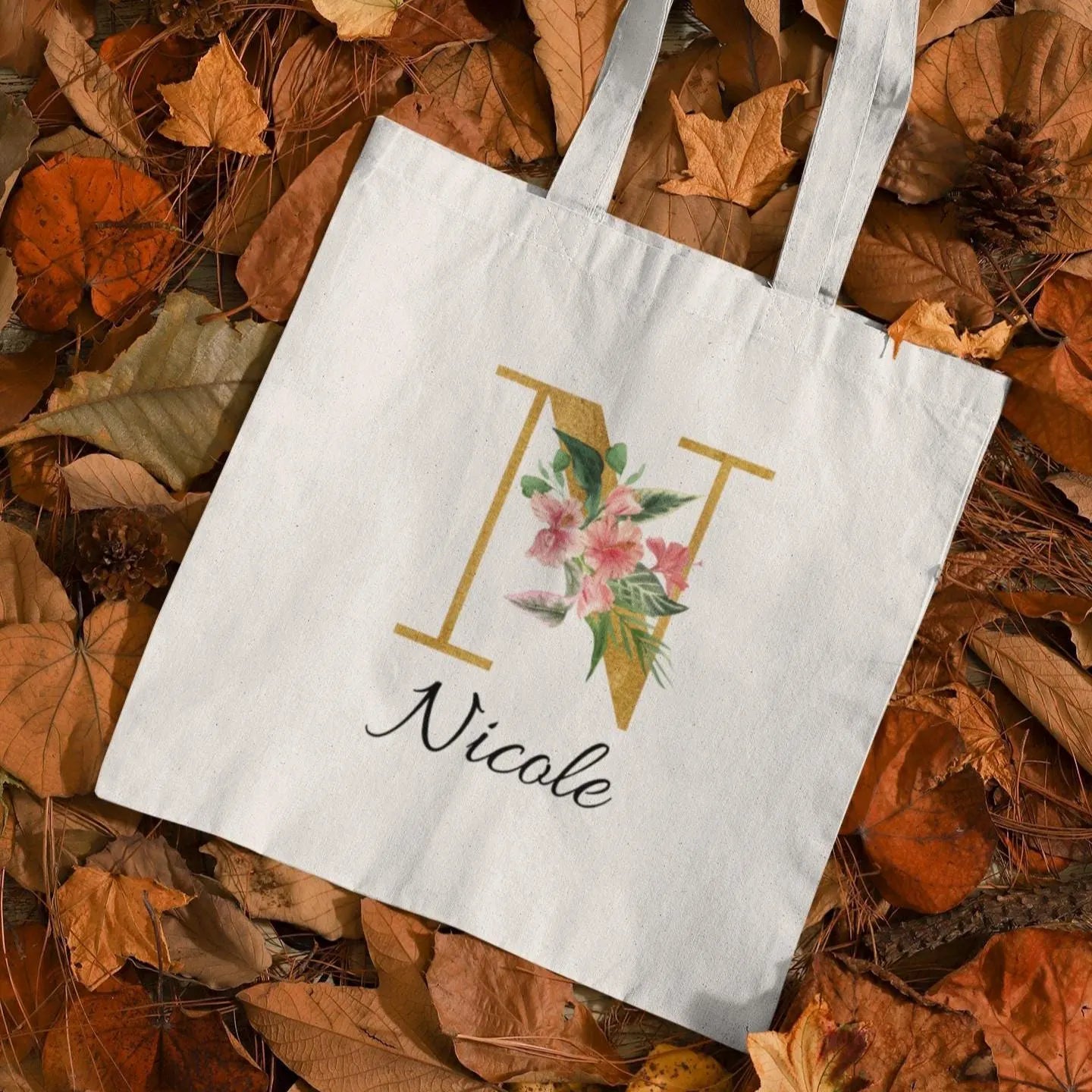 Hibiscus tropical floral custom tote bag with gold initial N and name Nicole, displayed on autumn leaves background
