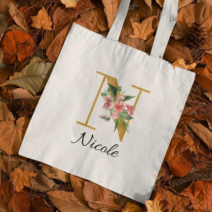 Hibiscus tropical floral custom tote bag with gold initial N and name Nicole, displayed on autumn leaves background