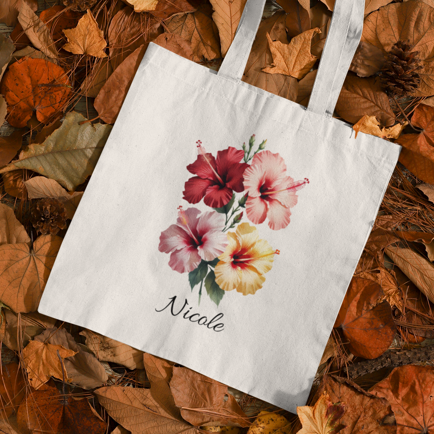 Tote bag with Hibiscus floral design and name 'Nicole' on a bed of autumn leaves