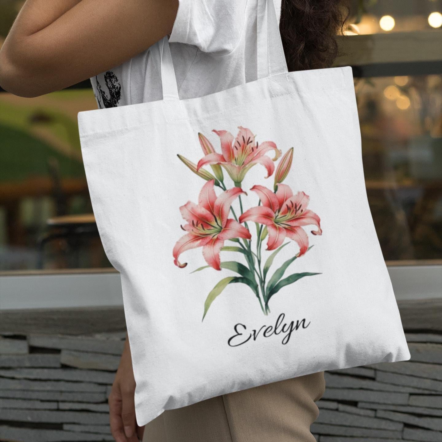 White tote bag with pink lilies and 'Evelyn' text held by a person outdoors.