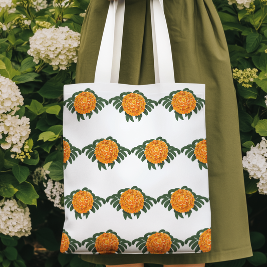 Marigold flower patterned tote bag with bright orange floral design held in a garden with white hydrangeas, showcasing bold botanical print on white fabric