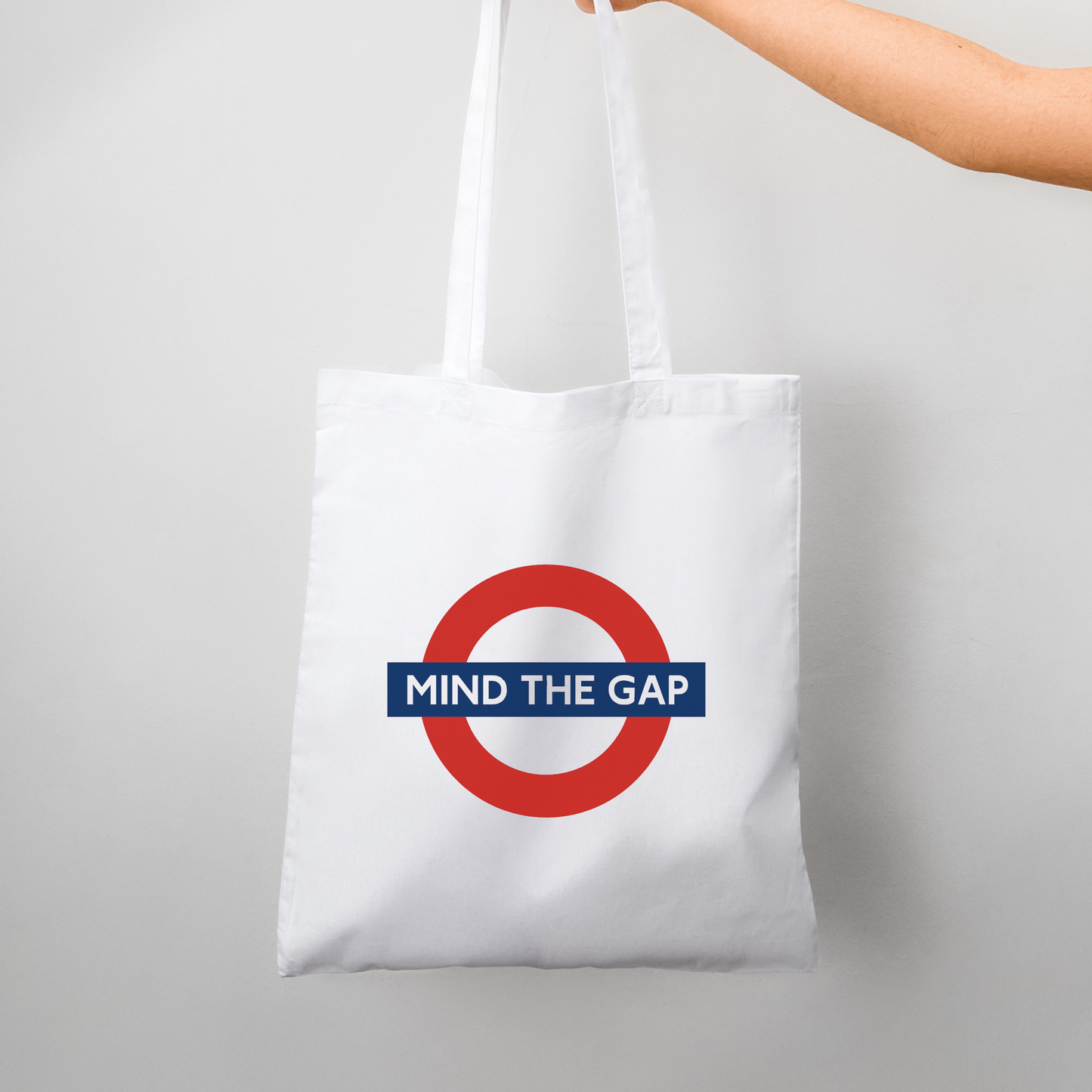 White cotton tote bag featuring the “Mind The Gap” London Underground design, held by hand – minimalist streetwear accessory