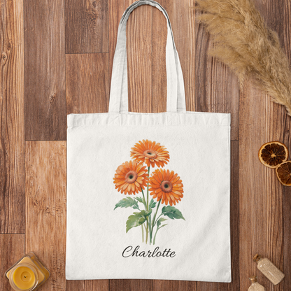 Personalised orange gerbera floral tote bag with name Charlotte printed, displayed on rustic wooden surface.