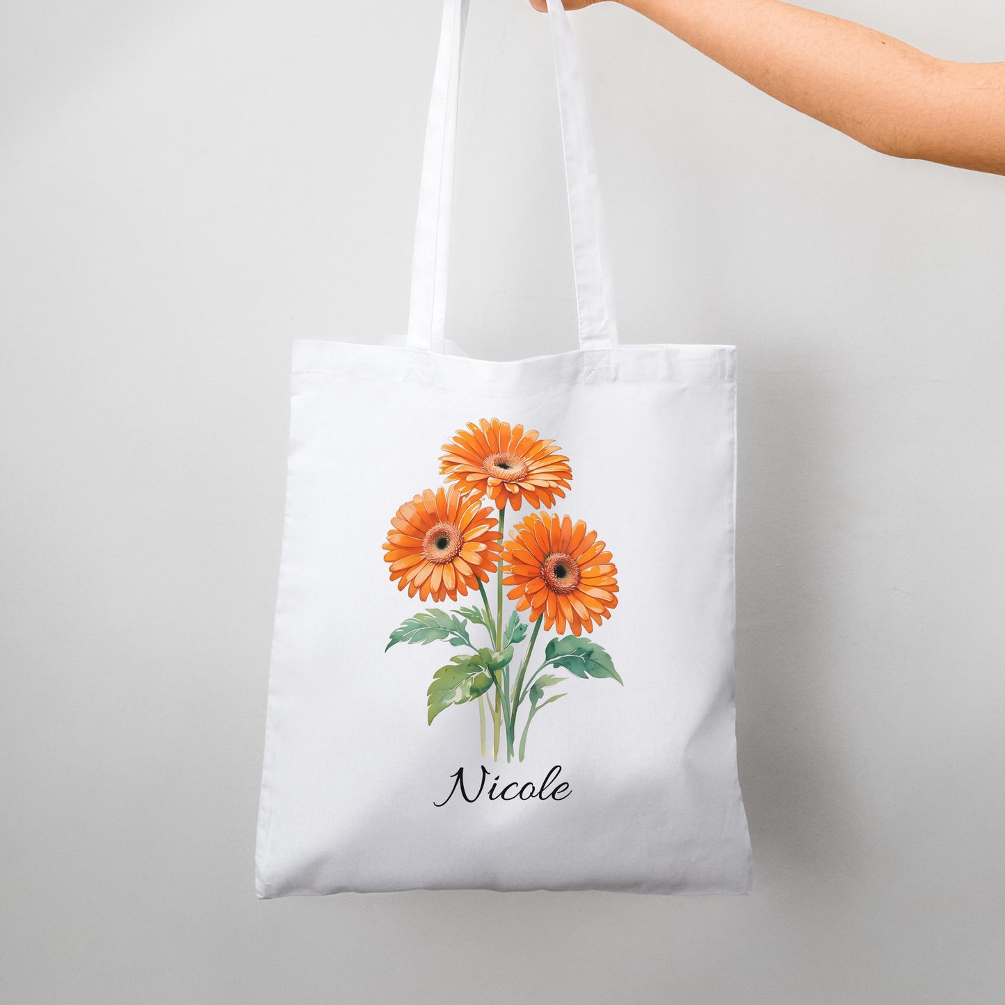 White tote bag with orange gerbera flower design and custom name “Nicole,” held by hand against neutral background