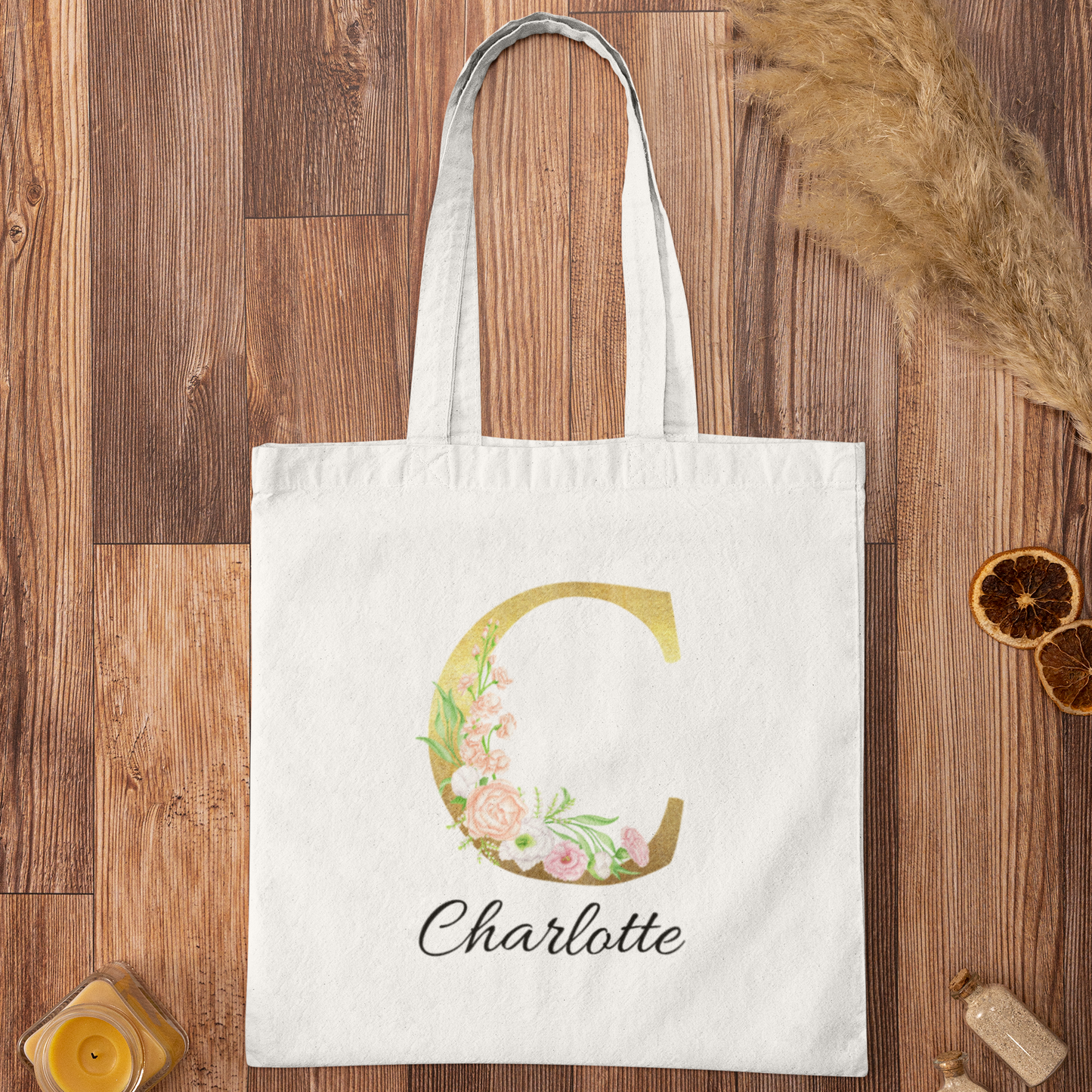 Personalized tote bag with gold letter C and pastel floral design, flatlay view on wooden background