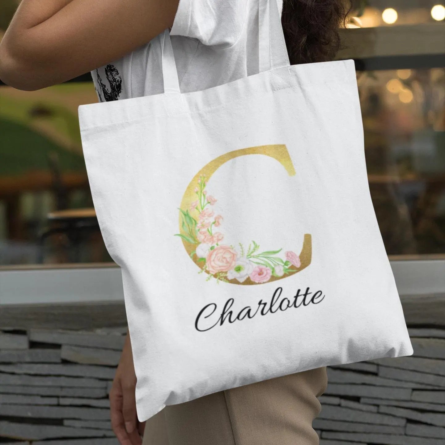 Gold floral initial tote bag with name Charlotte held by woman wearing casual outfit