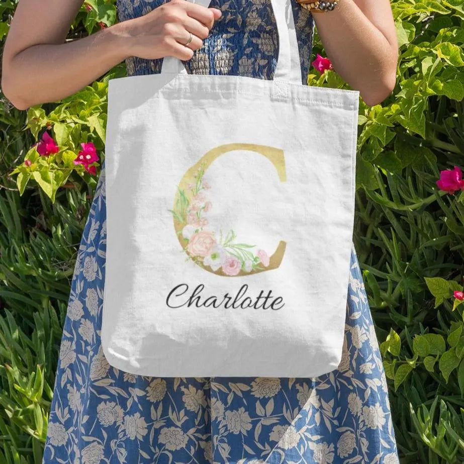 Gold and pastel floral initial tote bag with name Charlotte, held outdoors among greenery