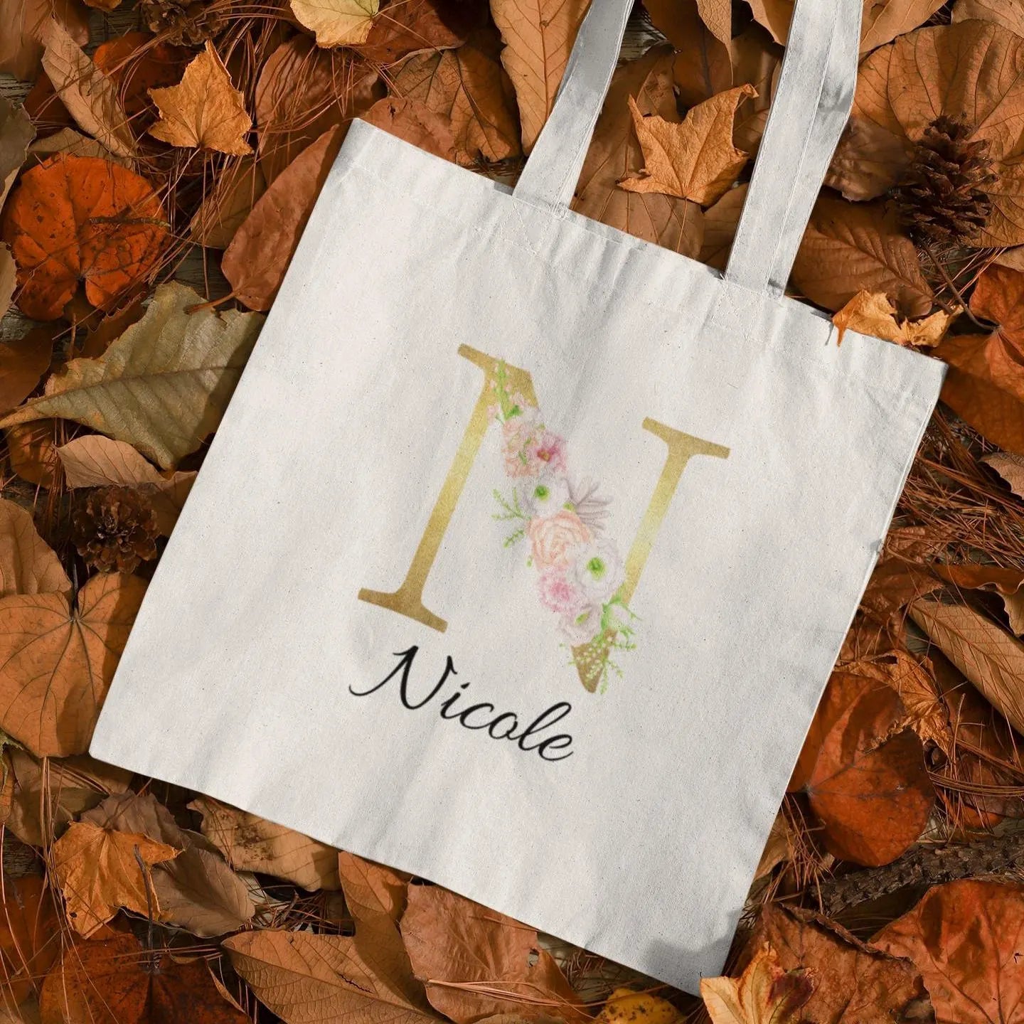 Tote bag with gold letter N and pastel flower design, flatlay on autumn leaves background