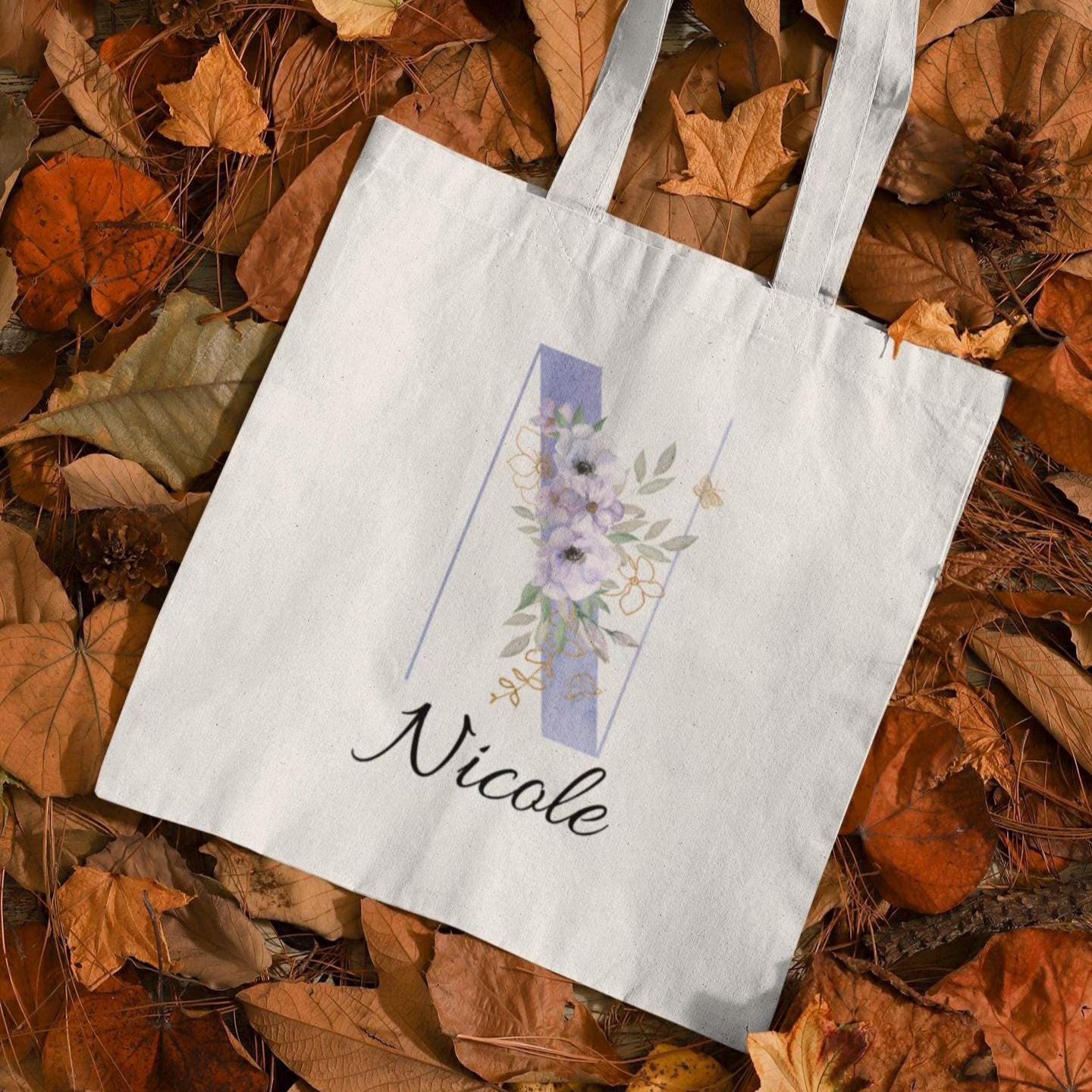 Pastel purple and cornflower blue floral custom tote bag with initial N and name Nicole, displayed on autumn leaves background with elegant anemone design