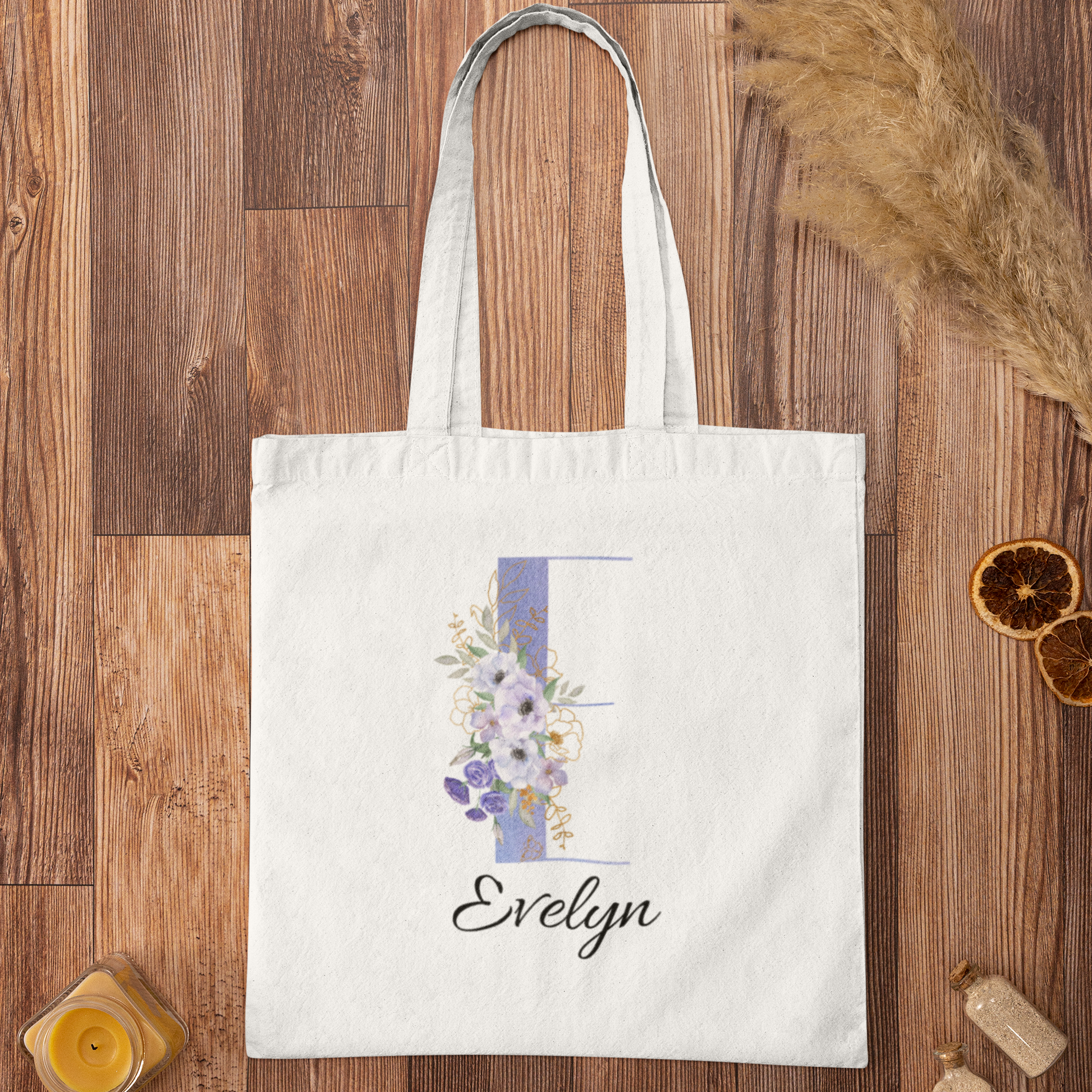 Flat lay of pastel purple and cornflower blue floral tote bag with initial letter E and name Evelyn, featuring soft anemone flowers on a white canvas bag