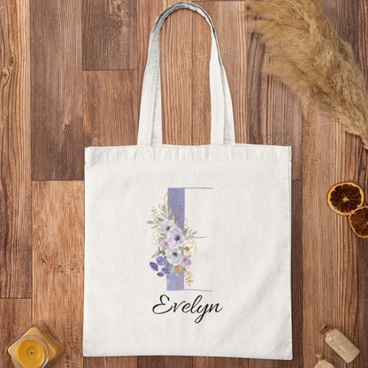 Flat lay of pastel purple and cornflower blue floral tote bag with initial letter E and name Evelyn, featuring soft anemone flowers on a white canvas bag