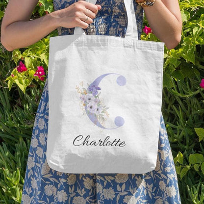 Woman outdoors holding pastel purple and cornflower blue floral tote bag with initial C and name Charlotte, featuring delicate anemone flower artwork on white fabric