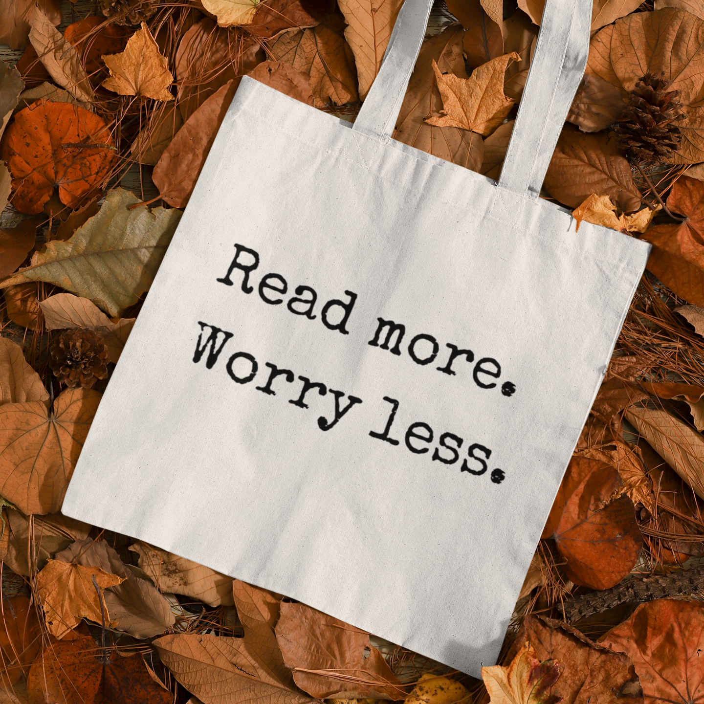 White minimalist tote bag with ‘Read more. Worry less.’ quote placed on autumn leaves – cozy reading aesthetic