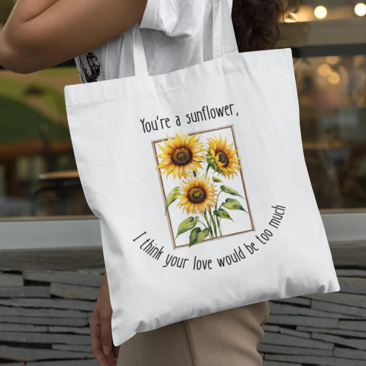 Tote bag with sunflower design and text held by a person outdoors.